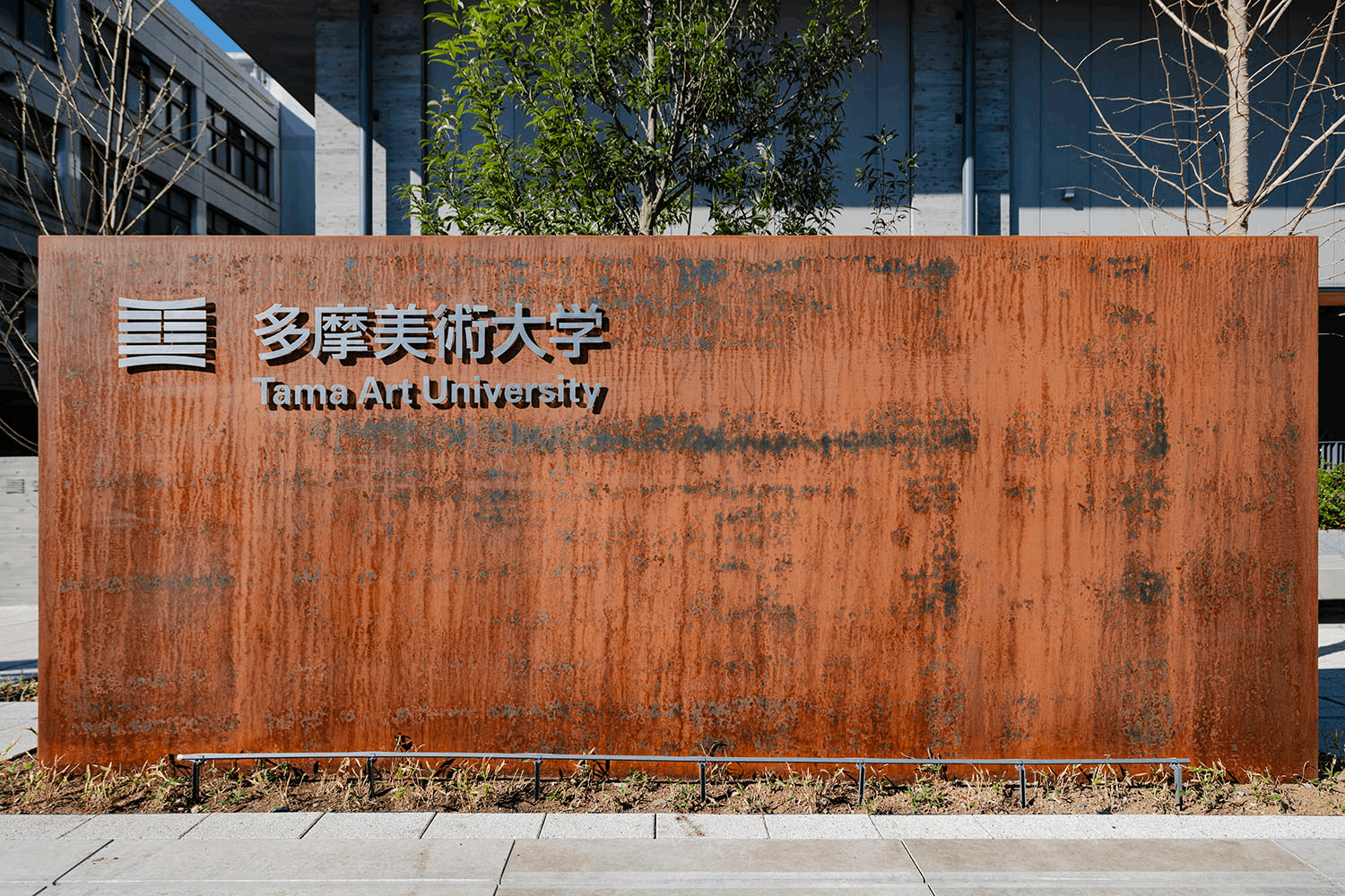 画像：多摩美術大学 上野毛キャンパス 大学銘板／正門の大学銘板。1970年前後、本学を中心に展開された「もの派」への敬意。無加工の鉄の素地が、その精神を今に伝える。